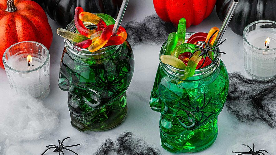 Zwei grüne Getränke in Totenkopf-Gläsern, dekoriert mit Gummibärchen und Spinnen, stehen auf einem Tisch. Im Hintergrund sind Kürbisse und Kerzen platziert. Die Szene hat ein gruseliges Halloween-Thema.