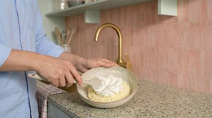 Hände einer Person rühren geschlagene Sahne in eine Schüssel mit Teig auf einer Küchenarbeitsplatte.