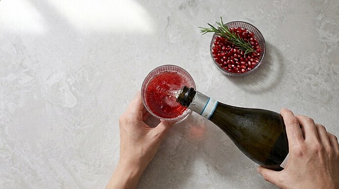Zwei Hände gießen Prosecco aus einer Flasche in ein Glas mit rotem Saft auf einer hellgrauen Arbeitsplatte.