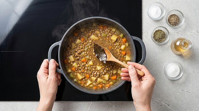 Dampfender Topf mit Linsensuppe und Würfeln aus Kartoffel und Karotte, ein Holzlöffel rührt ein Lorbeerblatt; Gewürzgläser daneben.