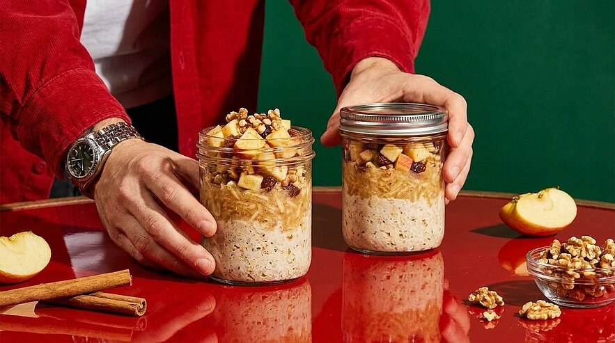 Zwei Einmachgläser mit Overnight-Oats, gekrönt von gewürfelten Äpfeln und Walnüssen; eine Hand setzt einen Deckel auf.