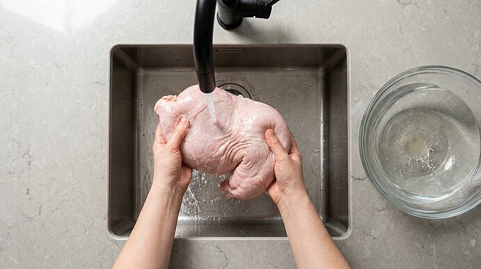 Hände halten ein rohes ganzes Hähnchen unter fließendem Wasser über einer Spüle; daneben eine Glasschüssel mit Wasser.