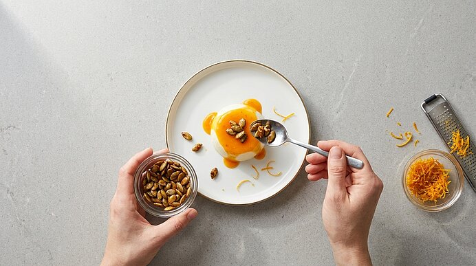 Hände garnieren auf einem weißen Teller eine kleine, runde weiße Creme mit orangener Sauce, Kürbiskernen und Orangenzesten; links ein Glas mit Kernen, rechts eine Schale mit Zesten und eine Reibe.