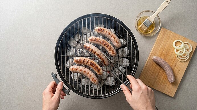 Ansicht von oben: Hände wenden gegrillte Bratwürste mit Zange auf rundem Holzkohlegrill; daneben Schneidebrett mit roher Bratwurst, Zwiebelringen und Schälchen mit Marinade.