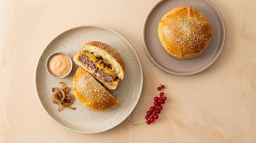 Zwei mit Sesam bestreute Brötchen, eines halbiert und gefüllt mit einem gebratenen Rinderpatty, geschmolzenem Käse und Gemüse, serviert auf Tellern mit einer Schale Sauce, karamellisierten Zwiebeln und roten Johannisbeeren.