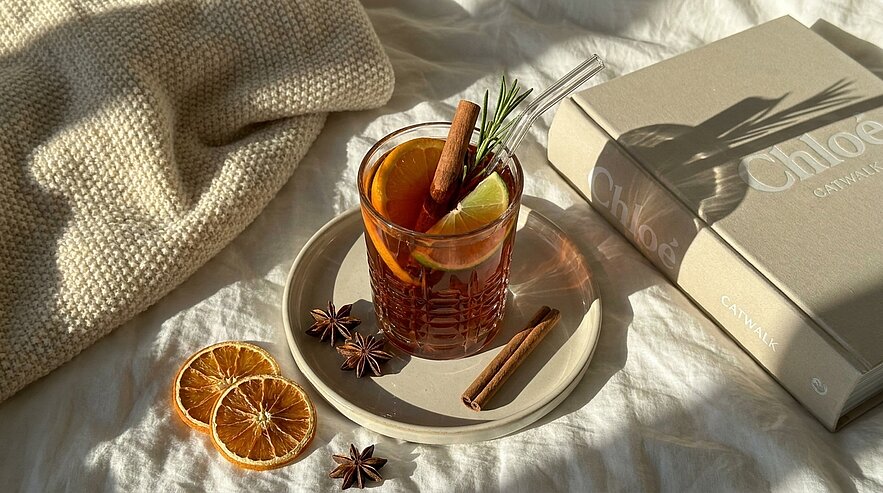 Glas mit warmem Gewürzgetränk auf Unterteller, garniert mit Zimtstange, Rosmarin und Zitronenscheibe; daneben getrocknete Orangenscheiben und Sternanis, vor einem Buch und kuscheliger Decke – gemütliche, herbstliche Szene.