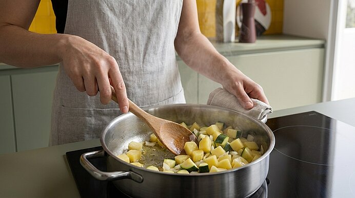 Person rührt gewürfelte Kartoffeln und Zucchini in einer Pfanne auf dem Herd mit einem Holzlöffel.