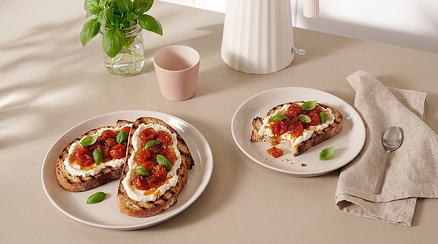 Geröstete Brotscheiben mit cremigem Frischkäse, karamellisierten Kirschtomaten und frischen Basilikumblättern, auf Tellern serviert.
