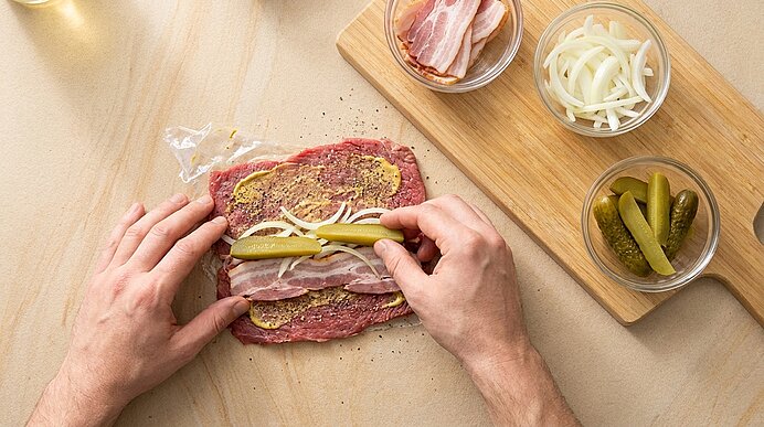 Zwei Hände belegen und rollen eine flache Rinderroulade mit Senf, Speckstreifen, Gewürzgurke und Zwiebelringen; kleine Schälchen mit Speck, Zwiebeln und Gurken stehen daneben.