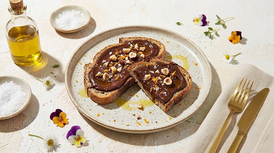 Zwei Scheiben Toastbrot auf einem Teller, bestrichen mit Schokoladen‑Haselnuss‑Aufstrich und bestreut mit gehackten Haselnüssen.