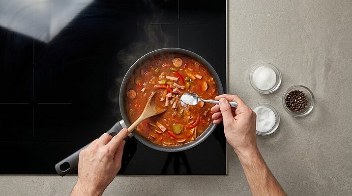 Hände rühren und würzen eine köchelnde, tomatenbasierte Gemüse‑Wurst‑Pfanne auf einer Herdplatte; Dampf steigt auf, daneben Schälchen mit Salz und Pfeffer.
