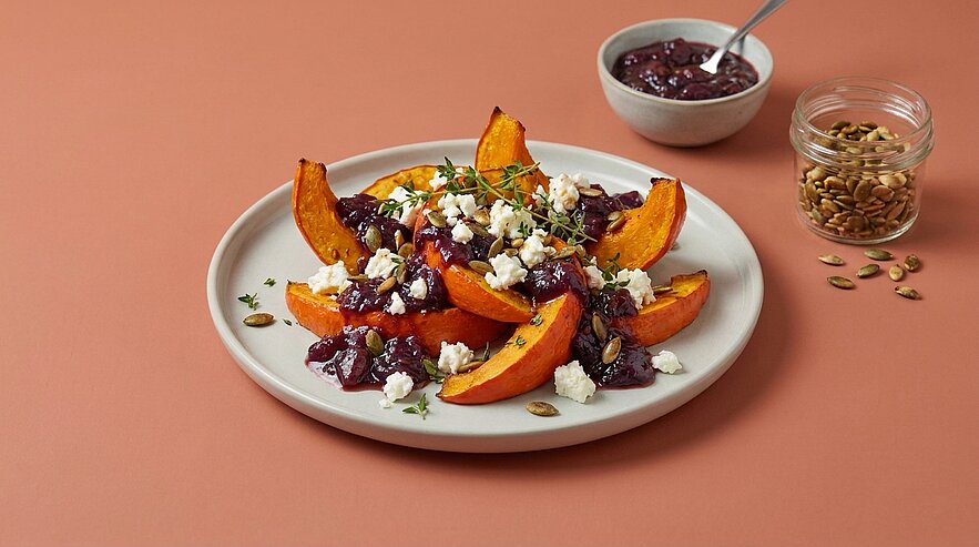 Teller mit gerösteten Kürbisspalten, garniert mit dunklem Beerenchutney, zerbröseltem Feta und frischen Kräutern.