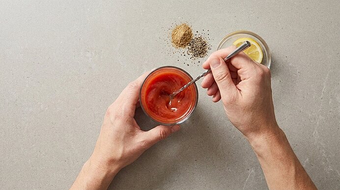 Zwei Hände rühren mit einem Löffel eine rote Sauce in einer Glasschale; daneben eine kleine Schale mit Zitronenscheiben und zwei Häufchen Gewürze auf hellem Untergrund.