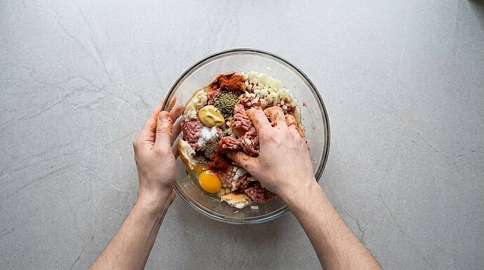 Zwei Hände vermengen in einer Glasschüssel Hackfleisch, ein rohes Ei, Semmelbrösel und Gewürze zu einer Fleischmasse.