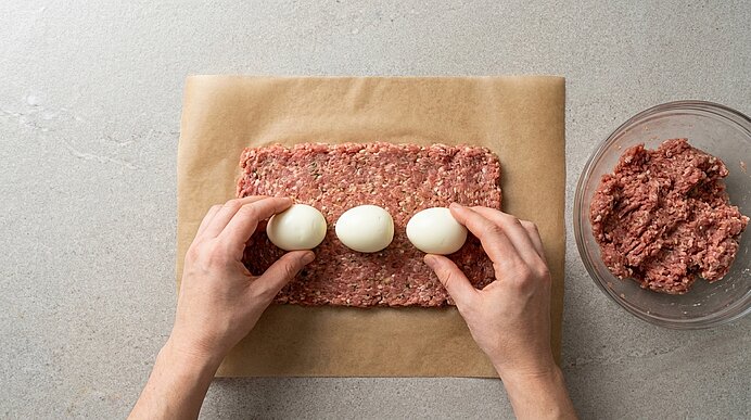Hände legen drei geschälte Eier in einer Reihe auf eine rechteckig ausgerollte Hackfleischmasse auf Backpapier; rechts eine Schüssel mit restlichem Hackfleisch.