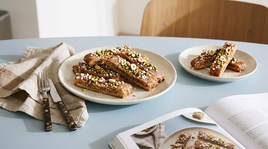 Teller mit schokoladenüberzogenen Biscotti-Stangen, garniert mit gehackten Nüssen und Zuckerstreuseln, auf einem Tisch neben Besteck und einem aufgeschlagenen Rezeptbuch.