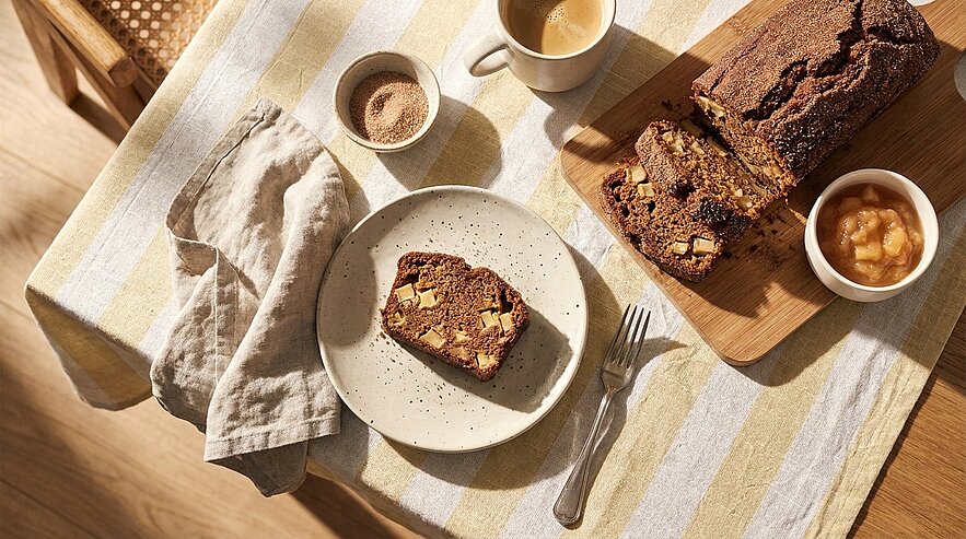 Auf einem Tisch: geschnittenes Nussbrot mit Nüssen auf einem Holzbrett, eine Scheibe auf einem Teller, Kaffeetasse, Schälchen mit Apfelmus, Gabel und Serviette.