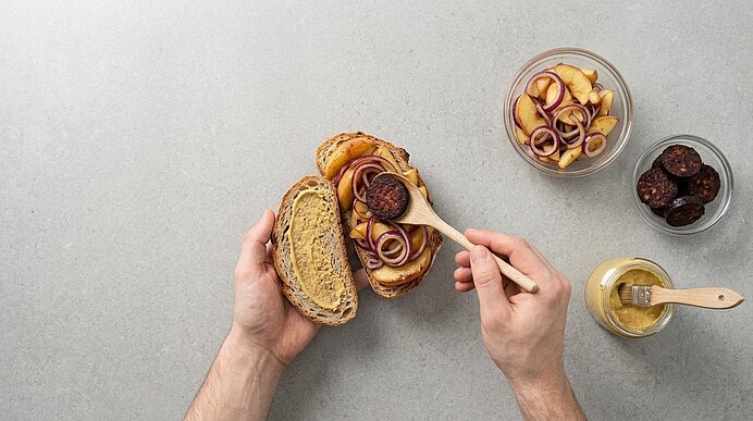 Zwei Hände legen gebratene Blutwurst mit einem Holzlöffel auf eine Brotscheibe mit Apfel-Zwiebel-Belag.