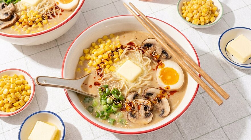 Schüssel cremiger Ramen: Nudeln in Brühe mit Mais, Champignons, halbiertem weichgekochtem Ei, Stück Butter, Frühlingszwiebeln und Chiliöl; Essstäbchen und Löffel.