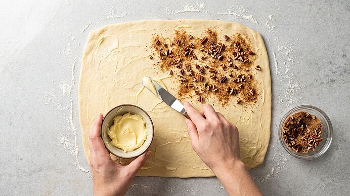 Hände bestreichen rechteckig ausgerollten Teig mit Butter; rechte Hälfte mit Zimt‑Zucker und gehackten Pekannüssen bestreut, kleine Schale mit Nüssen daneben.