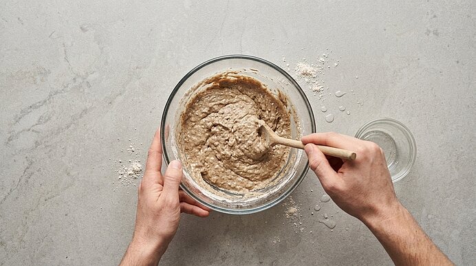 Zwei Hände verrühren weichen Roggensauerteig mit einem Holzlöffel in einer Glasschüssel auf hellgrauer Arbeitsplatte.