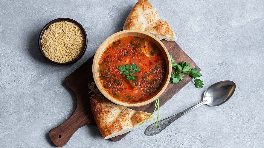 Eine Holzschüssel mit roter Suppe, garniert mit frischem Koriander, steht auf einem Holzbrett. Neben der Suppe liegen Stücke von knusprigem Brot und ein kleiner Schalen gefüllt mit ungekochten Reis. Der Hintergrund ist hell und schlicht.