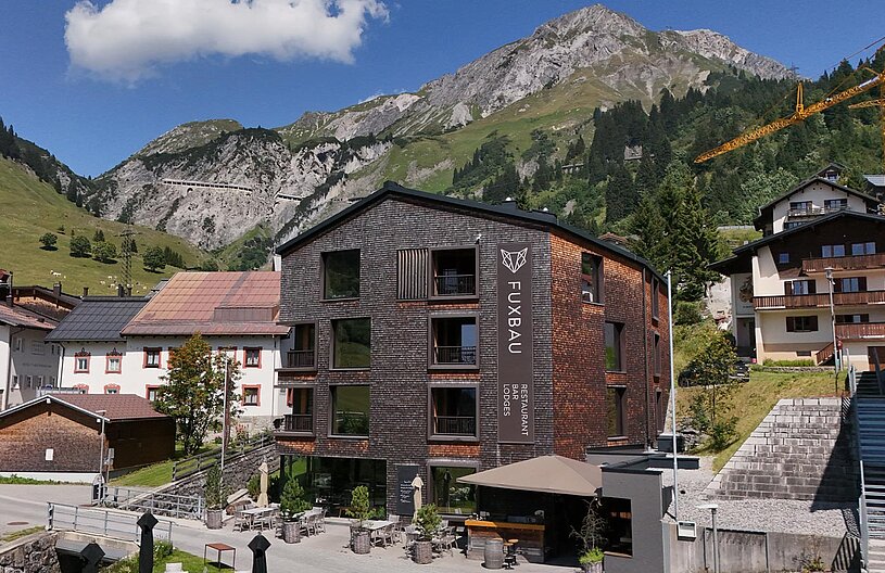 Ein modernes Hotelgebäude mit dem Namen „Fuxbau“ steht im Vordergrund, umgeben von Bergen und grünen Wiesen. Das Hotel hat große Fenster und eine Terrasse mit Sitzmöglichkeiten. Im Hintergrund sind Häuser und ein Kranausleger sichtbar, die die alpine Umgebung betonen.
