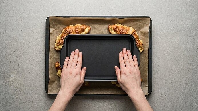 Zwei Hände drücken mit einer mattschwarzen Form gefüllte Croissants auf einem Backblech vorsichtig flach.