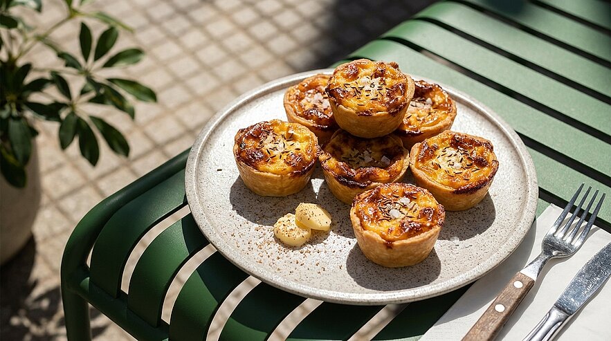 Sechs goldbraune Pastéis-de-Nata-Puddingtörtchen mit karamellisierter Oberfläche und Mandelblättchen auf einem hellen Keramikteller.