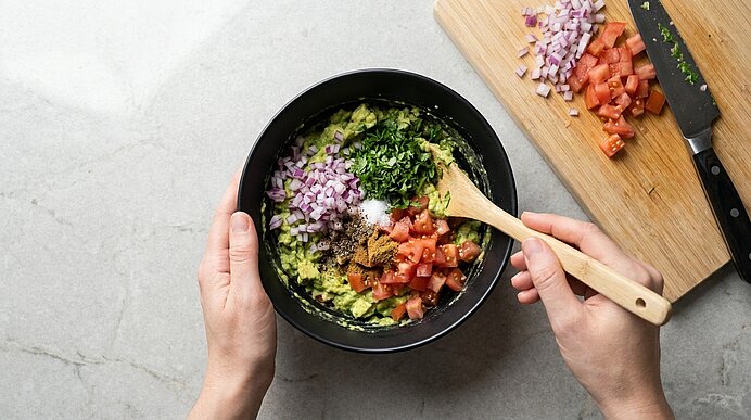 Zwei Hände heben gewürfelte Zwiebeln, Tomaten, Koriander und Gewürze mit Holzlöffel unter Guacamole.