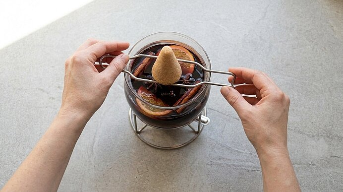 Zwei Hände halten eine Zange mit einem Zuckerhut über einem Glas Glühwein, in dem Orangenscheiben, Zimtstangen und Sternanis schwimmen.