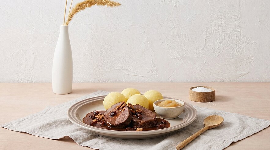 Teller mit in Scheiben geschnittenen Rinderrouladen in dunkler Soße, Kartoffelklößen und Apfelmus, daneben Salz und Holzlöffel.