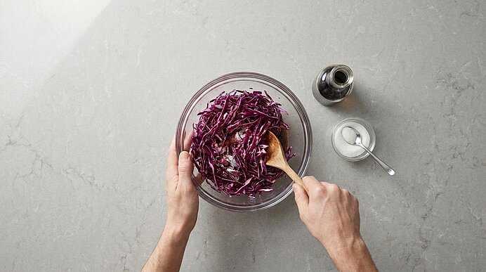 Zwei Hände mischen in einer Glasschüssel fein geschnittenen Rotkohl mit einem Holzlöffel.