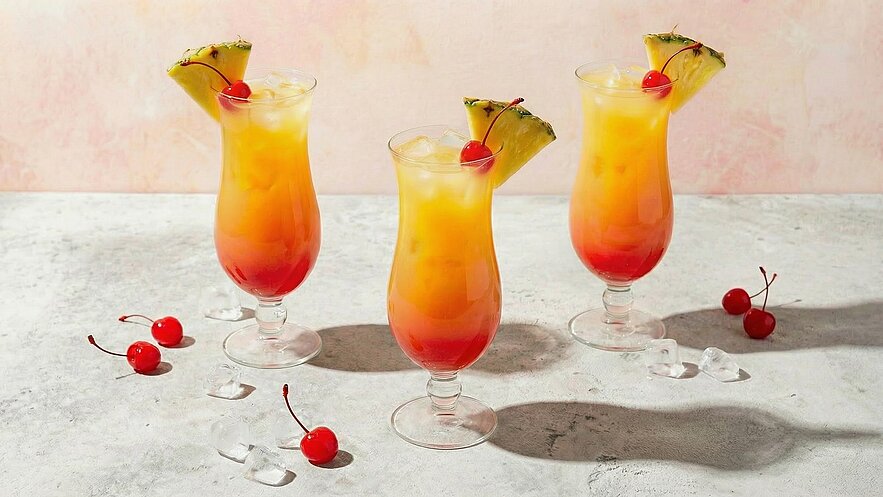 Drei Cocktails in hohen Gläsern stehen auf einem Tisch. Die Drinks haben eine leuchtend orange-gelbe Farbe mit roten Schichten am Boden. Jeder Drink ist mit einer Kirsche und einer Ananasstückchen dekoriert. Um die Gläser liegen einige Kirschen und Eiswürfel.