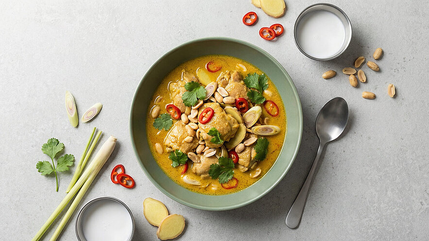 Eine grüne Schüssel mit cremiger Curry-Suppe, garniert mit Hühnerstücken, Erdnüssen, frischem Koriander und roten Chilis. Neben der Schüssel liegen frische Zutaten wie Zitronengras und Ingwer, sowie eine kleine Schale mit Kokosmilch.