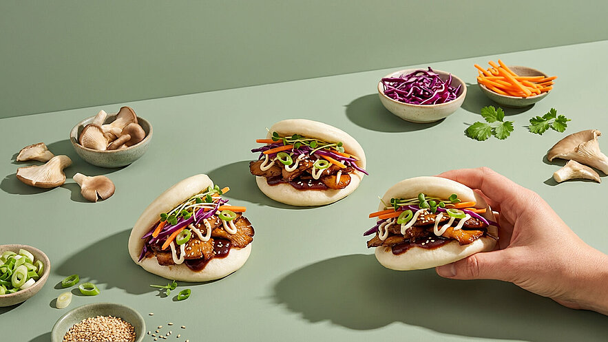 Drei gefüllte Bao-Brötchen liegen auf einem Tisch. Eines davon wird von einer Hand gehalten. Die Brötchen sind mit Hühnchen, buntem Gemüse und Saucen dekoriert. Im Hintergrund sind einige Schalen mit verschiedenen Zutaten wie Pilzen, Kohlgemüse und Karotten zu sehen.