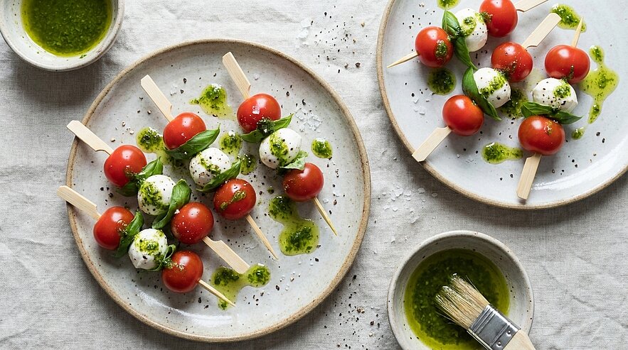 Drei Spieße mit Kirschtomaten und Mozzarella‑Kugeln, mit Basilikumpesto und schwarzem Pfeffer beträufelt, auf Tellern als Fingerfood angerichtet.