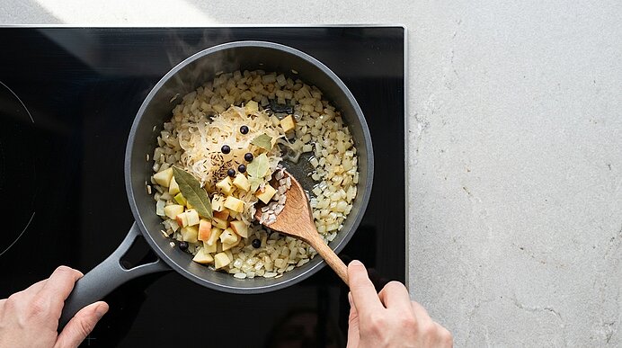 Zwei Hände rühren mit einem Holzlöffel in einem Topf auf dem Herd gewürfelte Zwiebeln und Äpfel mit Lorbeerblättern und Wacholderbeeren.