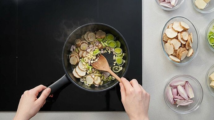 Zwei Hände dünsten Topinambur, Lauch, Schalotten und Knoblauch mit einem Holzlöffel in einem schwarzen Topf an.