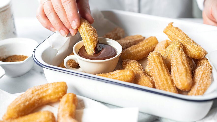 Eine Hand taucht ein goldbraunes Churro in eine kleine Schüssel mit Schokoladensoße. Im Hintergrund sind weitere Churros in einer weißen Schüssel zu sehen, die mit Zucker bestreut sind. Die Szene vermittelt Genuss und eine einladende Atmosphäre.