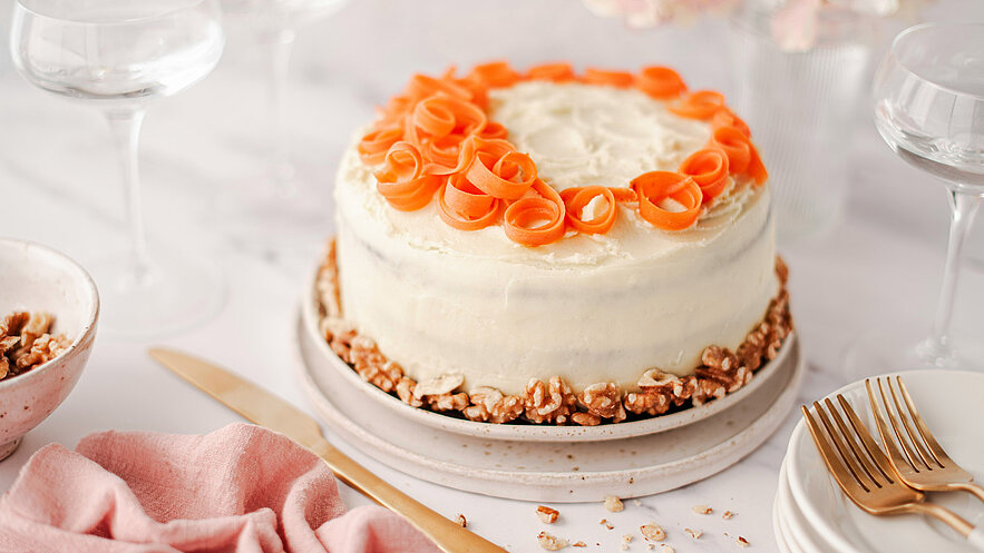 Eine dekorative Torte mit einer weißen Glasur und orangefarbenen Karotten-Röllchen oben drauf. Der Tortenteller ist mit einem Rand aus Nüssen verziert. Im Hintergrund sind eine rosa Serviette und Besteck sichtbar.