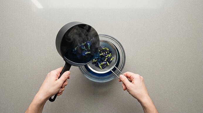 Zwei Hände sieben dampfende blaue Flüssigkeit mit blauen Blütenblättern aus einem kleinen Topf durch ein feines Sieb in eine Glasschüssel.