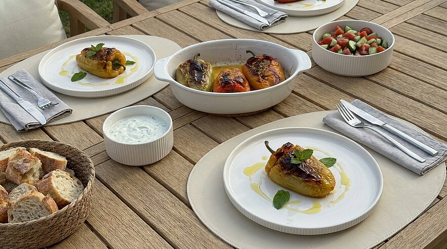 Der Tisch ist gedeckt mit mehreren Tellern, die gefüllte Paprika enthalten. In der Mitte steht eine Auflaufform mit weiteren gefüllten Paprika. Daneben befindet sich ein Schälchen mit Joghurt-Dip und ein Korb mit Brot. Im Hintergrund ist ein Salat aus Tomaten und Gurken zu sehen.