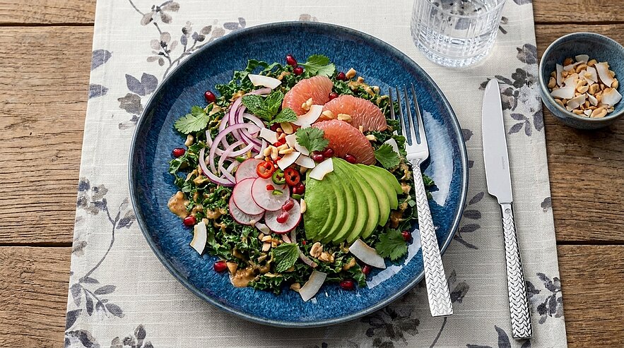 Auf dem Bild ist ein bunter Salat auf einem blauen Teller zu sehen. Der Salat besteht aus Blattgemüse, Radieschen, Avocado, Grapefruit, Zwiebeln und Kräutern. Neben dem Teller liegen Besteck und ein Glas Wasser. Im Hintergrund sieht man einen kleinen Schüssel mit Kokosraspeln.