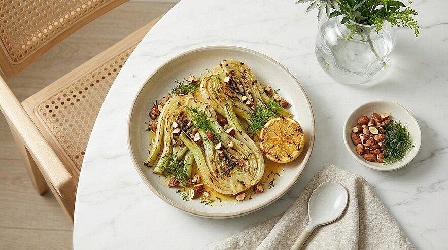 Ein Teller mit gegrilltem Gemüse, darunter Zucchini und Fenchel, garniert mit gerösteten Mandeln und frischem Dill. Ein halbierter, gegrillter Zitrone liegt daneben. Im Hintergrund ist eine schlichte, helle Tischoberfläche und eine Pflanzenvase zu sehen.
