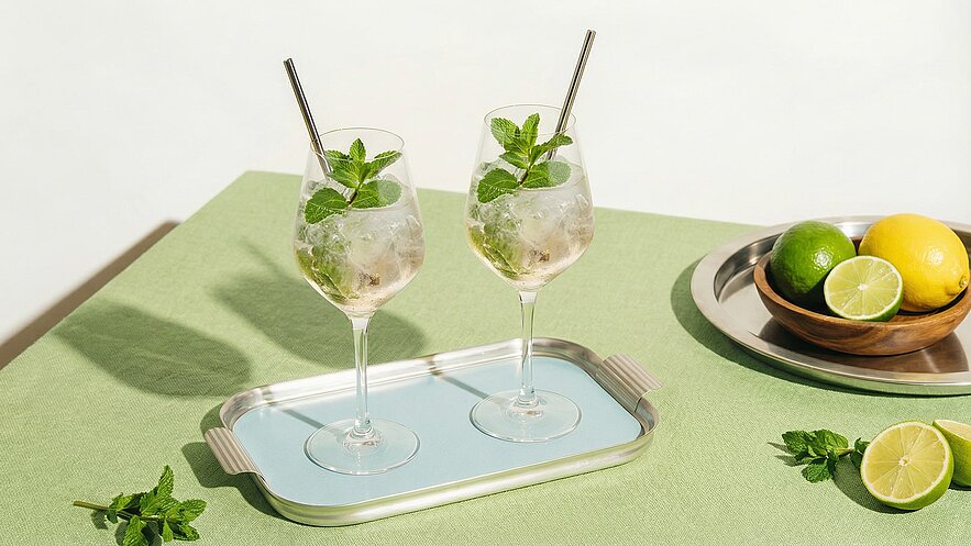 Zwei Gläser mit erfrischendem Getränk, gefüllt mit Eis und frischer Minze, stehen auf einem blauen Tablett. Im Hintergrund liegen eine Schale mit Zitronen und Limetten sowie frische Minzblätter auf einem grünen Tisch.