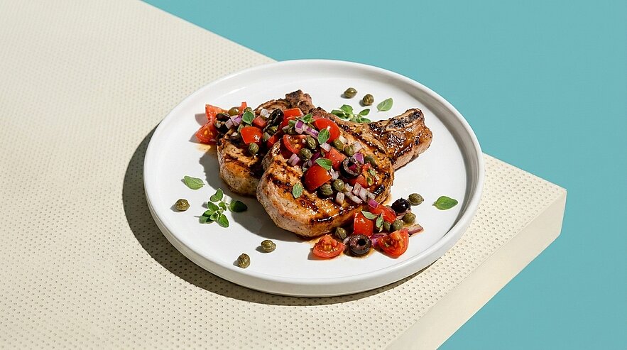 Zwei gegrillte Koteletts auf weißem Teller, garniert mit Tomaten-Oliven-Salsa, Kapern und frischen Kräutern.