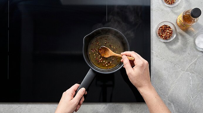 Zwei Hände halten einen kleinen schwarzen Topf auf einem Kochfeld, in dem mit einem Holzlöffel eine würzige Sauce verrührt wird; Dampf steigt auf. Rechts stehen Schälchen mit Chiliflocken und eine Flasche Öl.