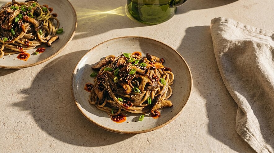 Teller mit gebratenen Udon-ähnlichen Nudeln und sautierten Pilzen, beträufelt mit Chili-Öl, bestreut mit Sesam und Frühlingszwiebeln.