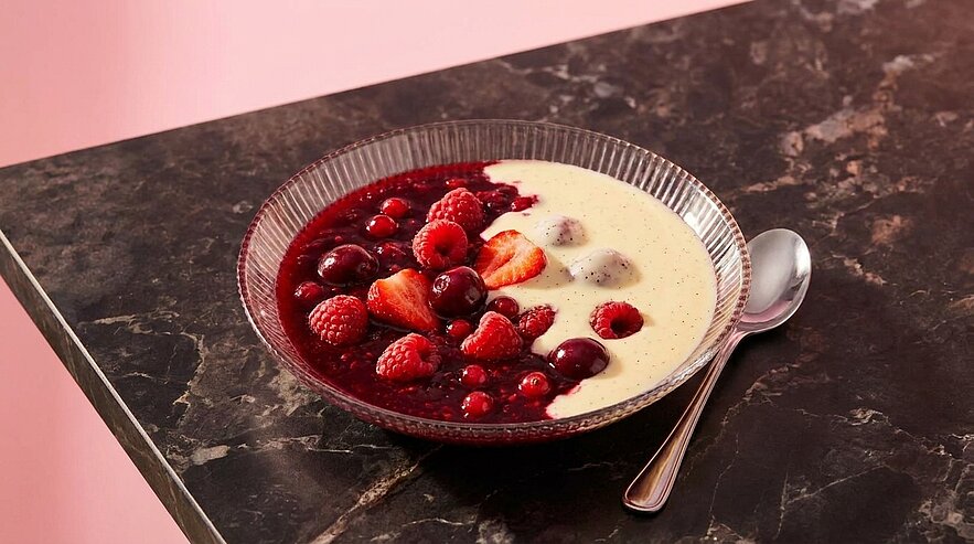 Glasschale mit cremiger Vanillecreme, darauf rote Beeren (Himbeeren, Erdbeeren, Kirschen) und Beerensoße; ein Silberlöffel liegt daneben.